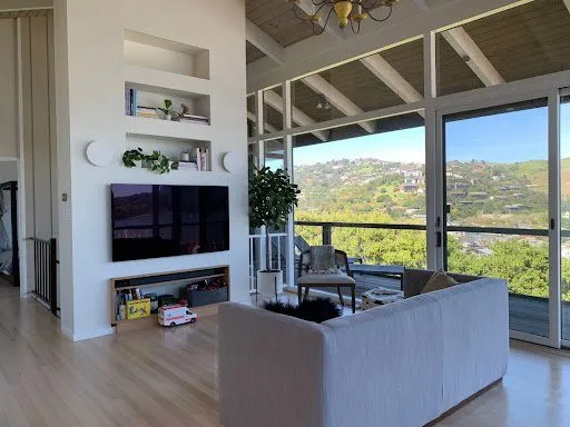 Existing living room before renovation: TV wall with built-in niches, grey sofa and sliding glass doors to the deck with hillside views