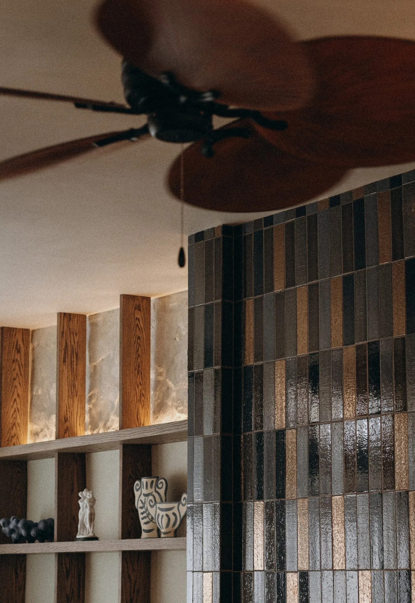 Palm-blade ceiling fan over a glazed wall of vertical tiles in black and bronze, backlit stone niches with ceramics beyond — Alisa: Wine and Friends - restaurant interior design