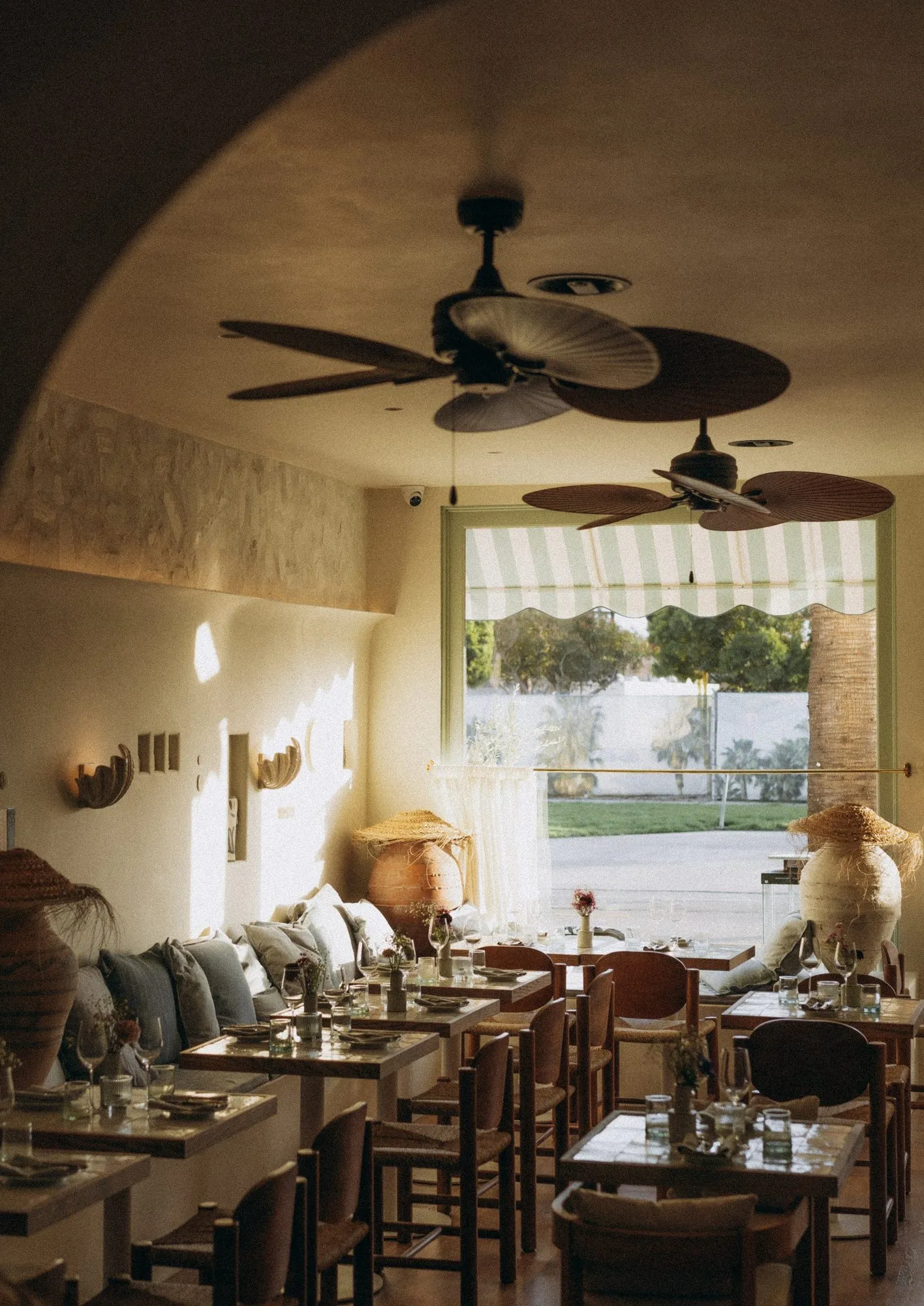 Sunlit dining room with tiled tables, wicker-seat chairs, palm ceiling fans and a striped scalloped awning at the window — Alisa: Wine and Friends - restaurant interior design