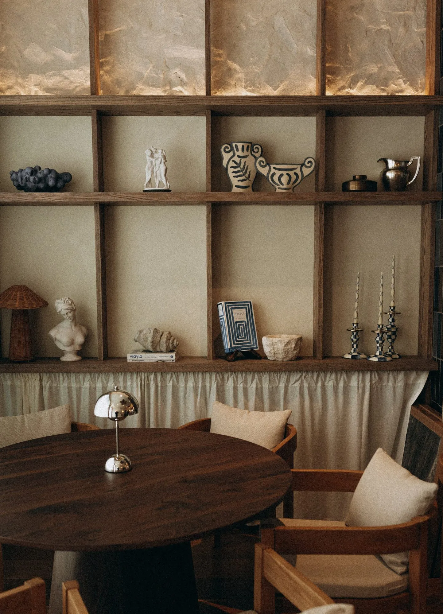Oak display grid with Greek-patterned vases, plaster bust and candlesticks over a skirted banquette and round walnut table — Alisa: Wine and Friends - restaurant interior design