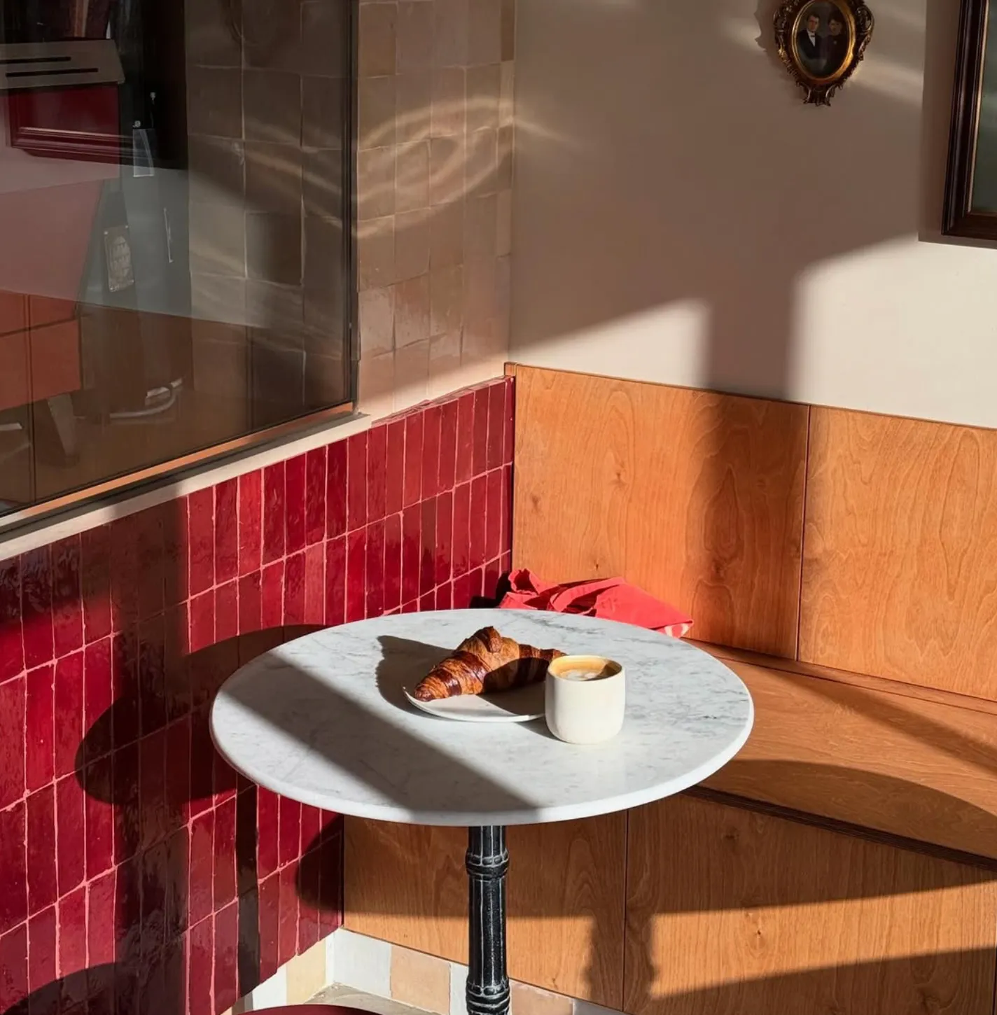 Rendering: cafe corner with a round marble table, croissant and coffee, burgundy tile wainscot, oak bench and sunlit window