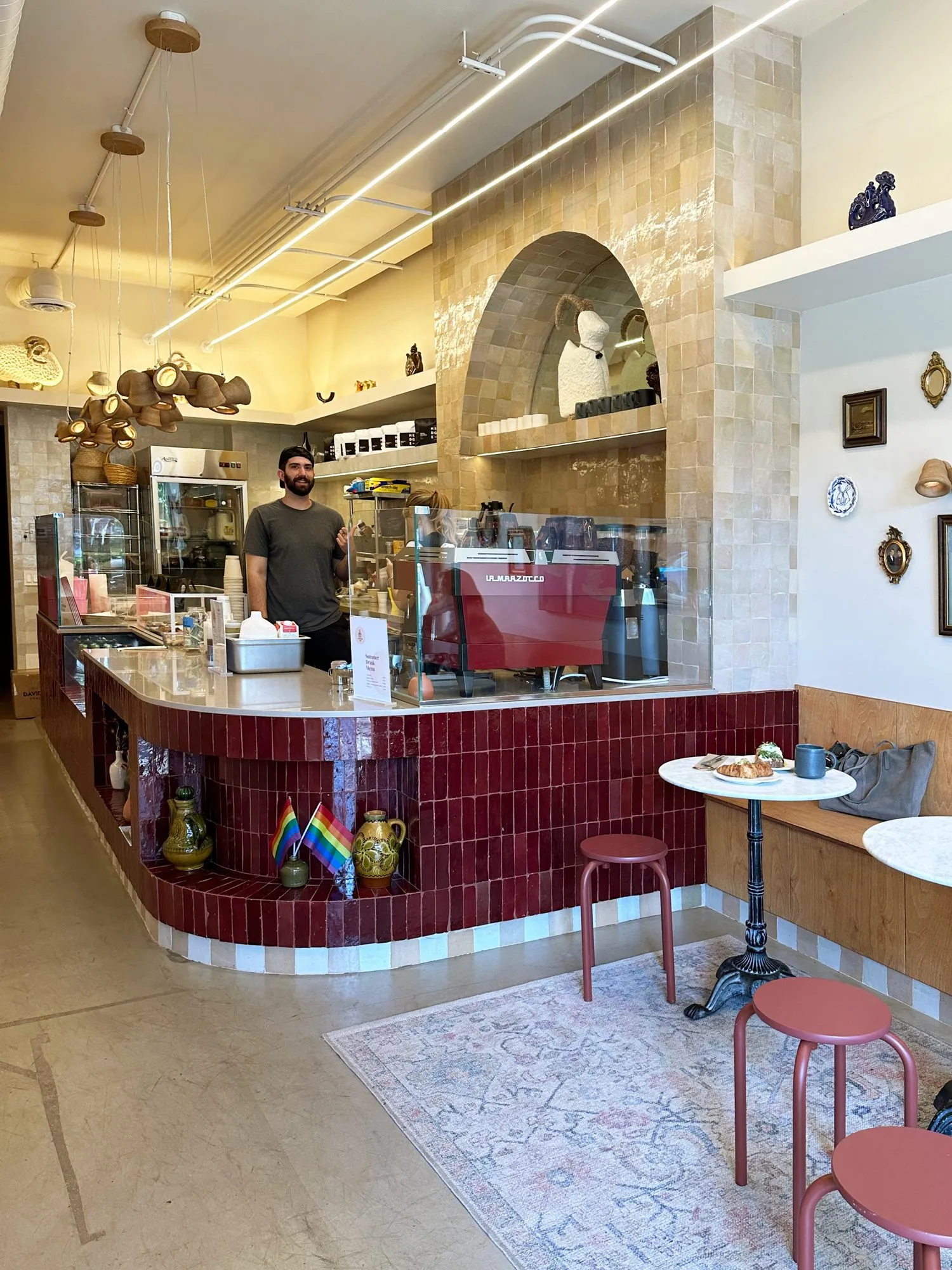 Finished bakery interior: curved burgundy-tiled counter with red espresso machine, barista, arched zellige wall and pink stools