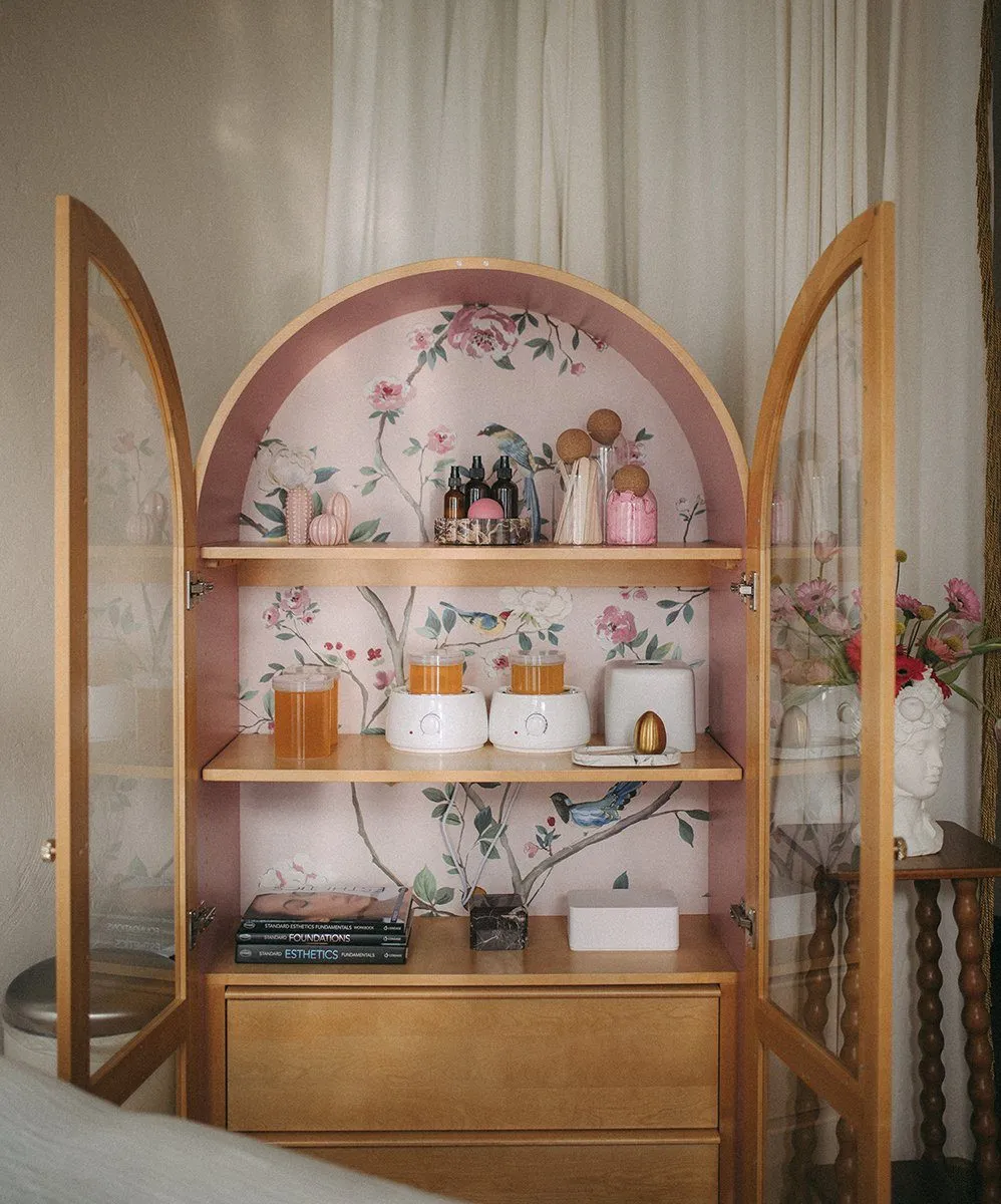 Arched maple cabinet lined with pink chinoiserie wallpaper, wax warmers, honey jar and esthetics books on its shelves — Relovi - wellness sugaring studio interior design