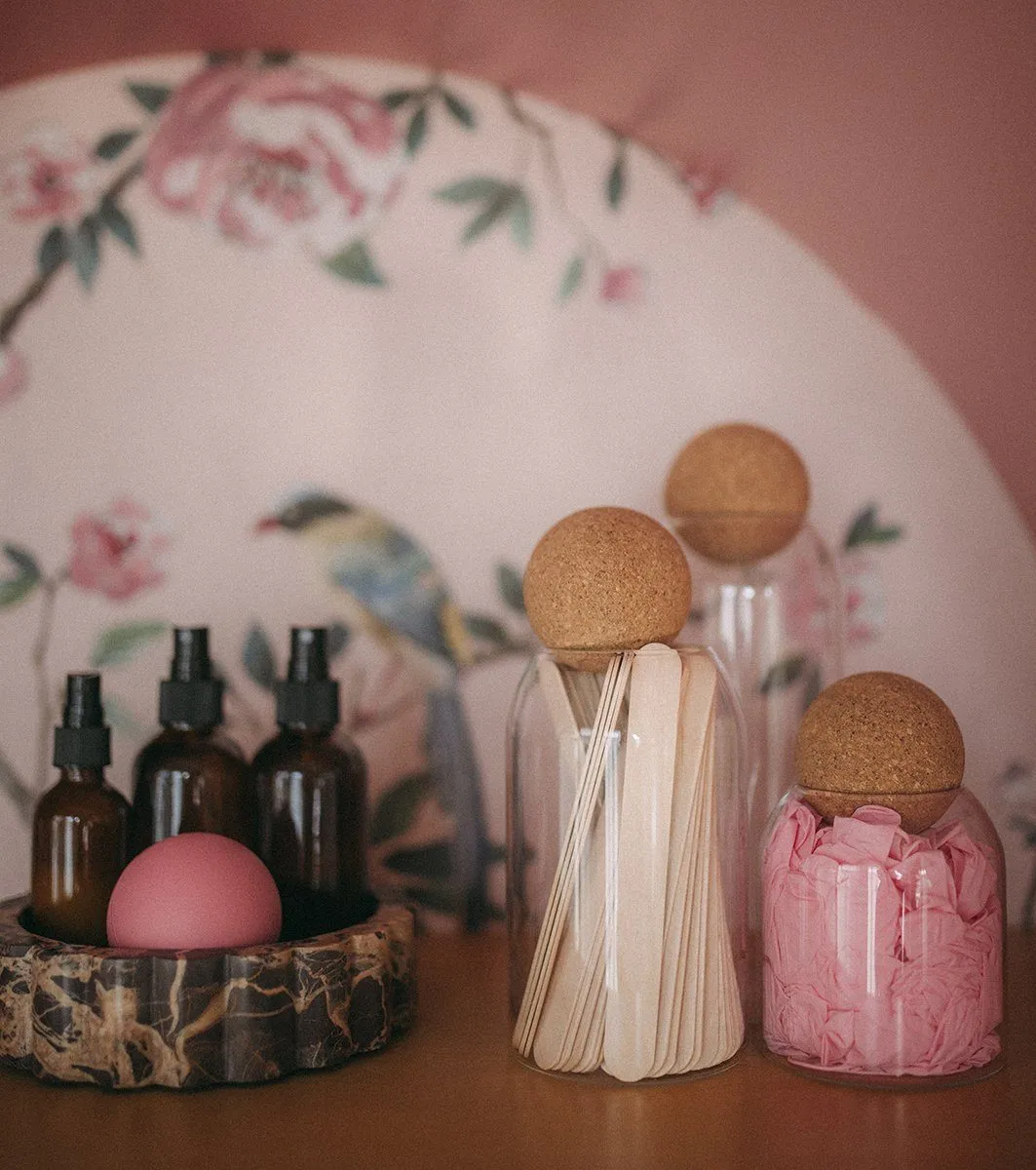 Amber spray bottles in a scalloped marble bowl beside cork-topped jars of wooden applicator sticks, floral chinoiserie behind — Relovi - wellness sugaring studio interior design