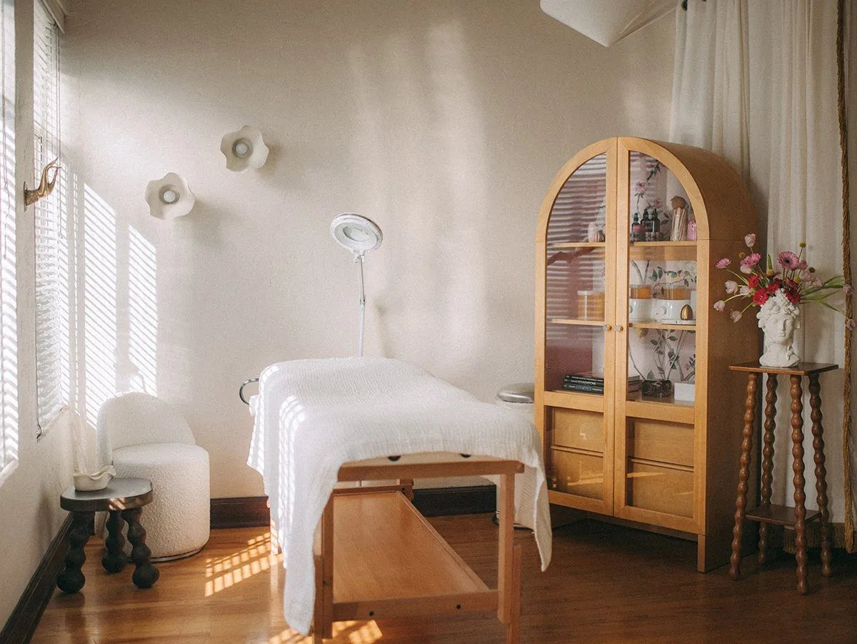 Treatment room with draped massage table, arched glass-front maple cabinet of products, magnifying lamp and white bouclé chair — Relovi - wellness sugaring studio interior design