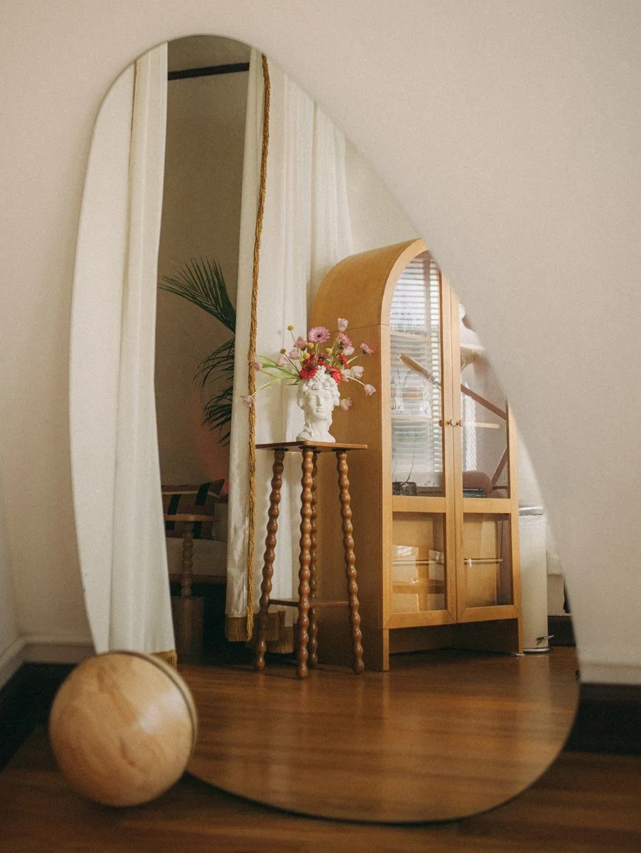 Organic-shaped floor mirror reflecting a bobbin side table with flower-crowned bust vase and the arched display cabinet — Relovi - wellness sugaring studio interior design