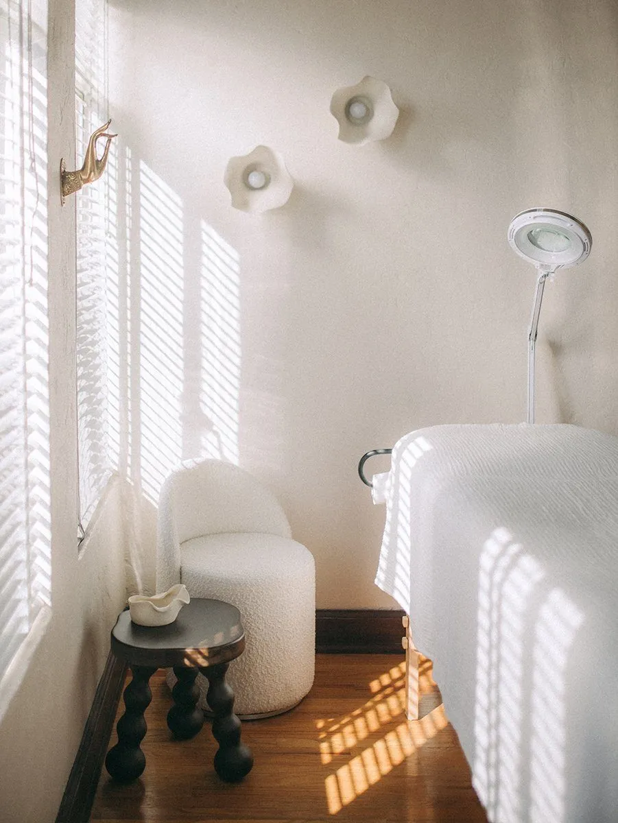 Corner with white bouclé slipper chair, black bobbin stool and plaster flower sconces in striped light through the blinds — Relovi - wellness sugaring studio interior design