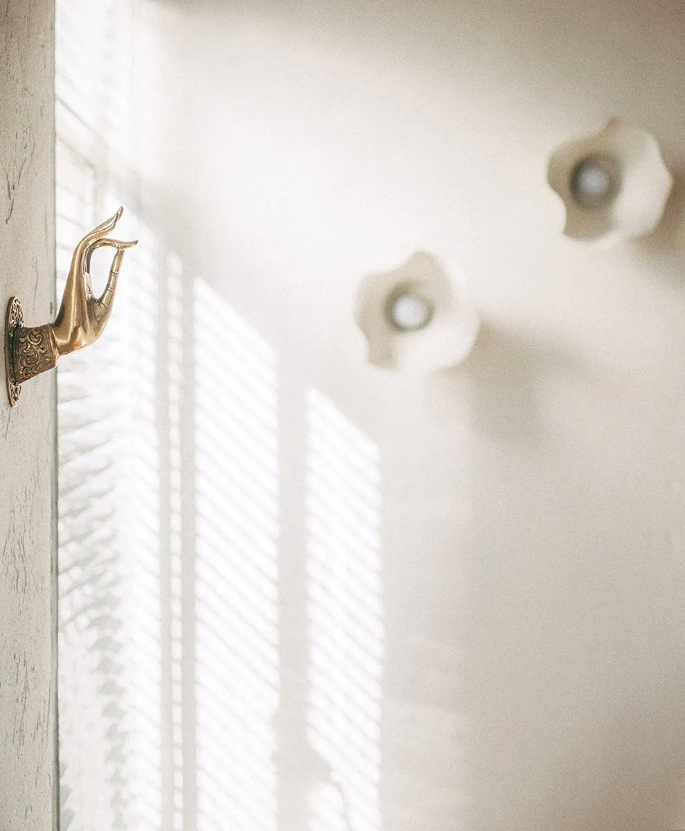 Brass mudra-hand wall hook beside the window, two flower-shaped plaster sconces in soft focus beyond — Relovi - wellness sugaring studio interior design