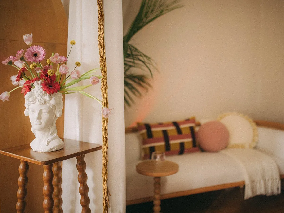 Classical bust vase of pink tulips and gerberas on a bobbin pedestal, fringed curtain and cream settee soft-focused beyond — Relovi - wellness sugaring studio interior design
