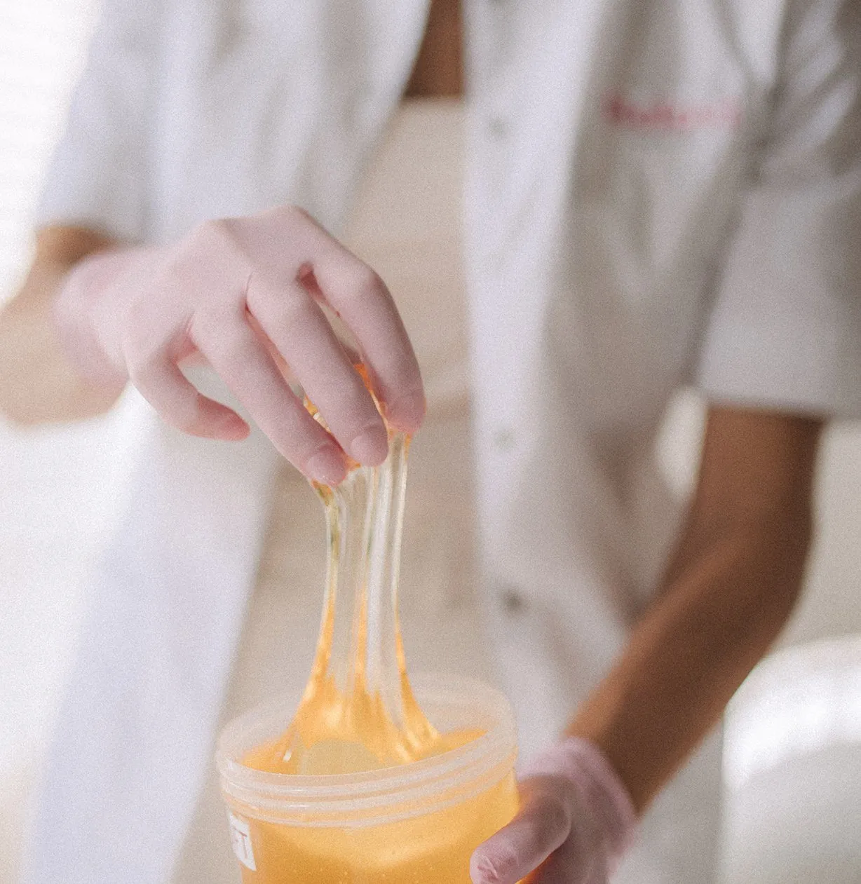 Close-up of hands stretching golden sugaring paste from its jar, practitioner in a white uniform behind — Relovi - wellness sugaring studio interior design