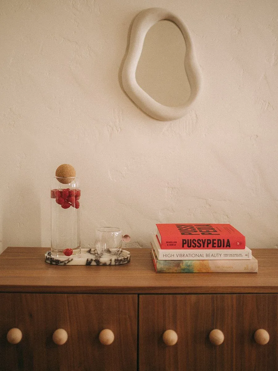 Walnut sideboard with wooden ball knobs, stacked wellness books, cork-ball carafe of raspberry water and a wavy plaster mirror — Relovi - wellness sugaring studio interior design