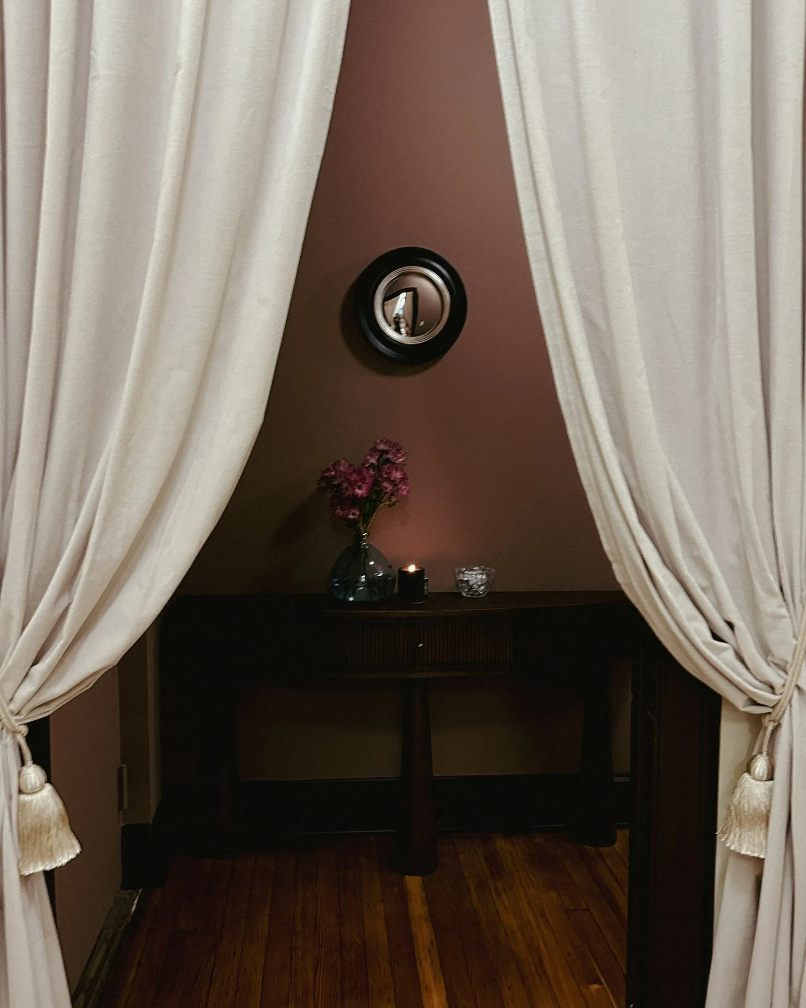 Tasseled linen curtains parting to a mauve alcove with convex mirror, dark console, fresh flowers and a lit candle — The Aesthetic Era - Medical SPA interior design